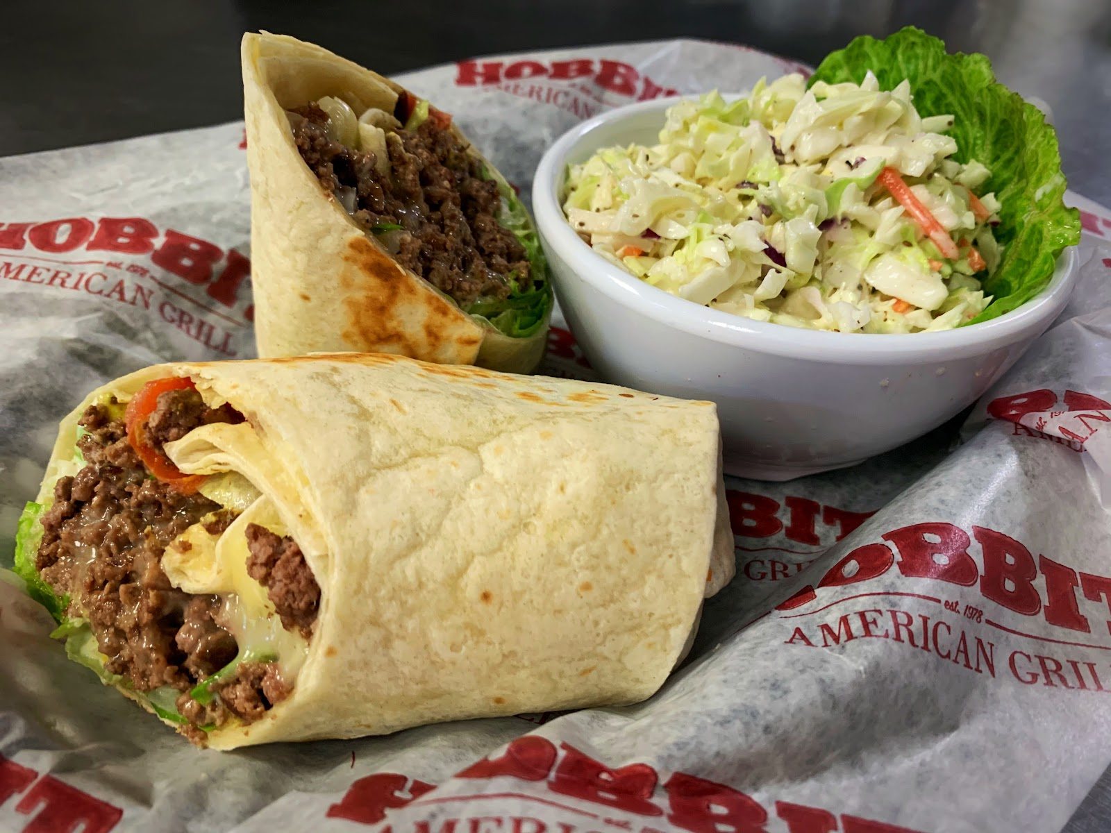 A wrap cut in half beside a bowl of coleslaw, served on Hobbit American Grill paper.