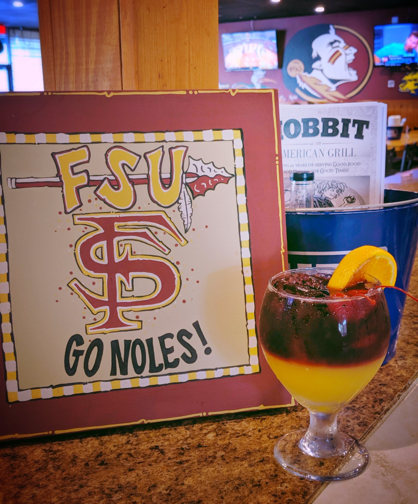 A hand-lettered FSU “Go Noles” sign on the bar beside a tall layered garnet and gold drink.