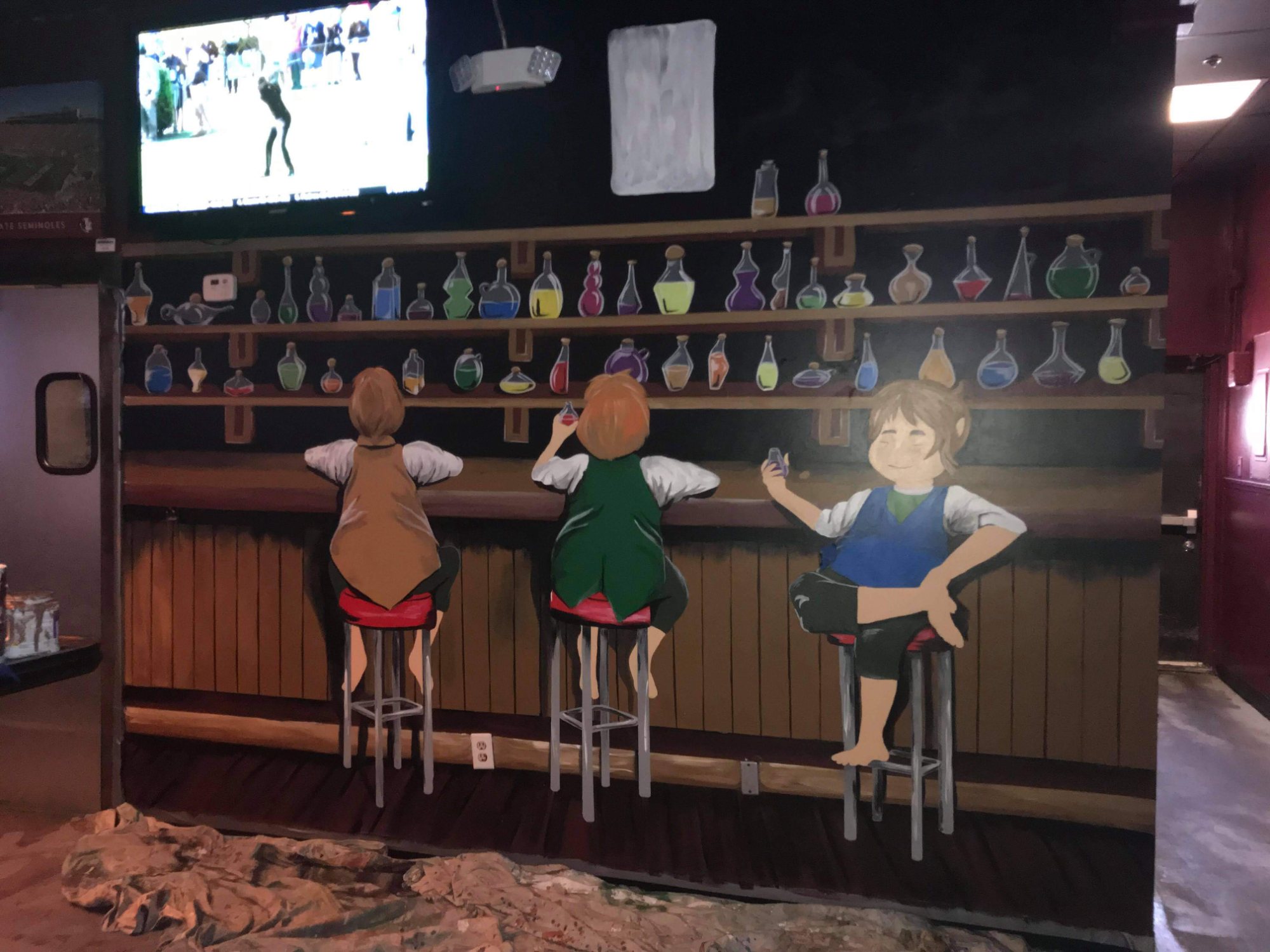 The hand-painted mural behind the bar: three hobbits on barstools facing shelves of coloured bottles, with TVs above.