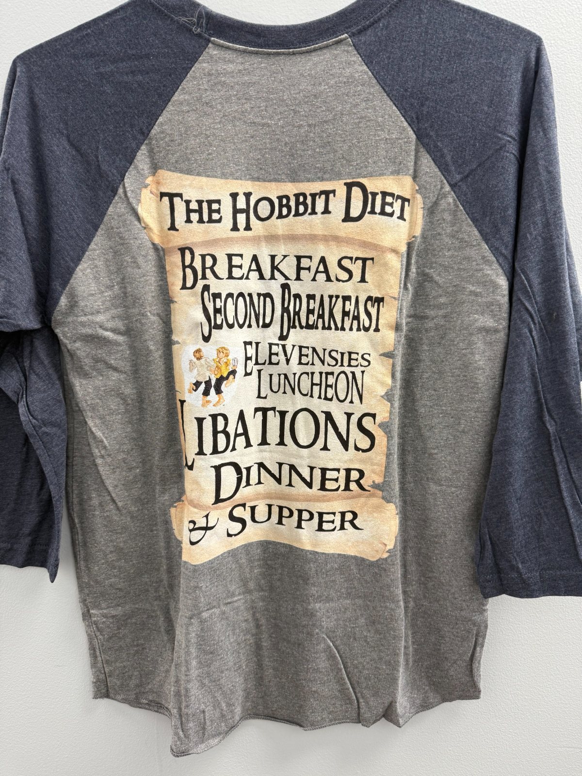 The back of a grey and navy baseball shirt printed with a scroll reading The Hobbit Diet: Breakfast, Second Breakfast, Elevensies, Luncheon, Libations, Dinner and Supper.
