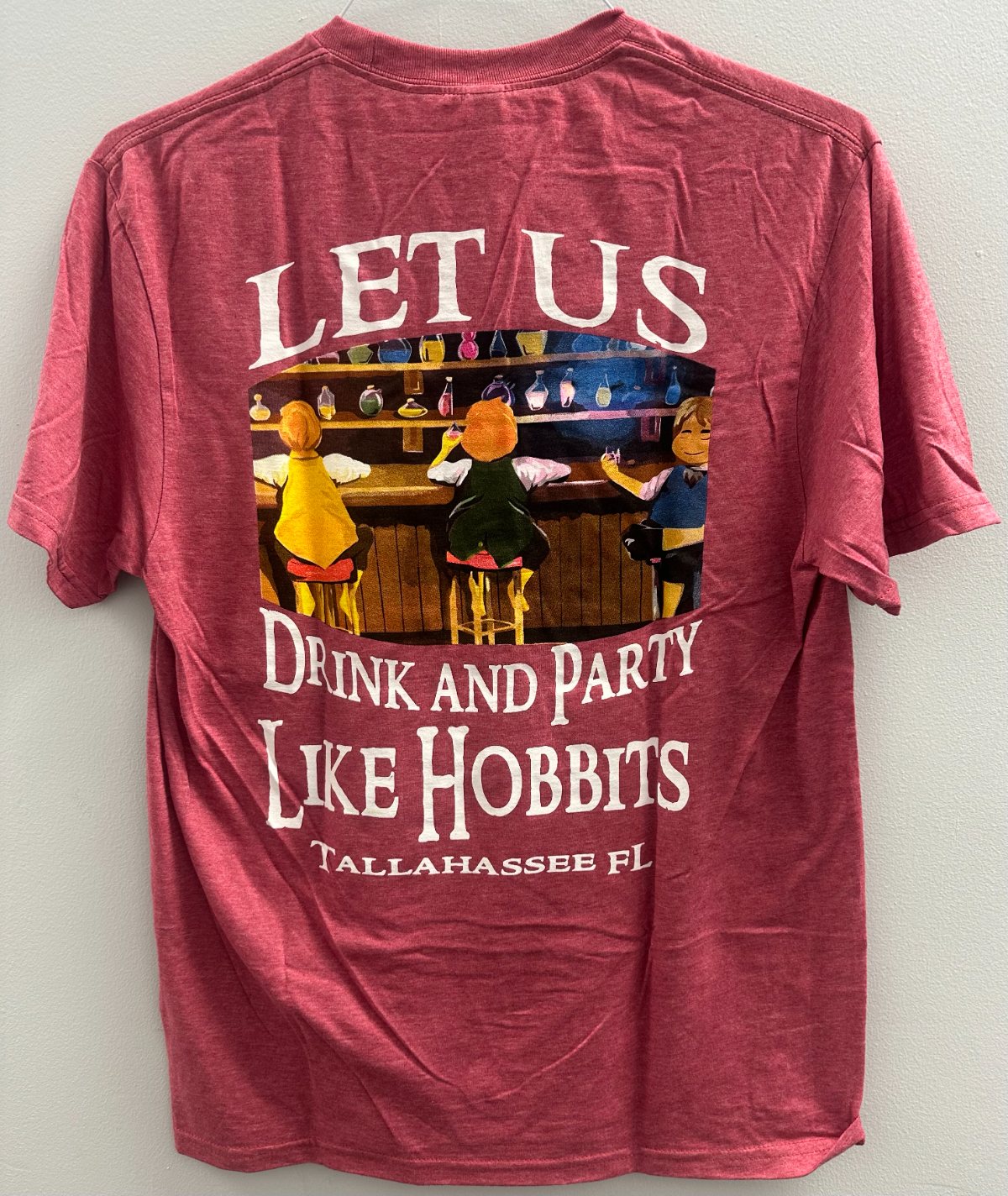 A heather garnet T-shirt reading Let Us Drink and Party Like Hobbits, Tallahassee FL, over a drawing of hobbits at a bar.